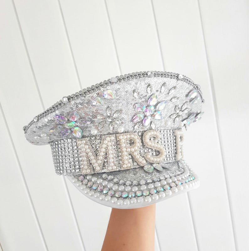 Op de afbeelding: Zilveren pailletten hoed met het woord "MRS" in parelachtige kralen. De hoed is versierd met iriserende strass-steentjes en parels. De hoed heeft een klep en is waarschijnlijk een bruidsaccessoire.