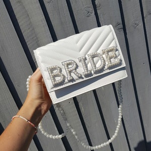 May include: White quilted clutch bag with a pearl embellished "BRIDE" lettering. The bag has a pearl chain strap.