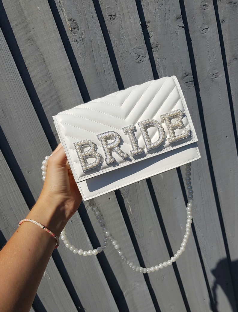 May include: White quilted clutch bag with a pearl embellished "BRIDE" lettering. The bag has a pearl chain strap.