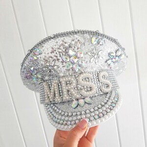 Op de afbeelding: Een zilveren en witte decoratieve hoed met het woord "MRS" gespeld in grote parelletters. De hoed is bedekt met pailletten, strass-steentjes en parels, met een decoratief bloemmotief erop.