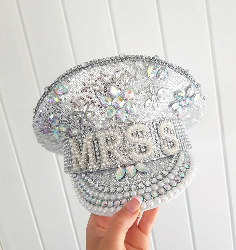 Op de afbeelding: Een zilveren en witte decoratieve hoed met het woord "MRS" gespeld in grote parelletters. De hoed is bedekt met pailletten, strass-steentjes en parels, met een decoratief bloemmotief erop.