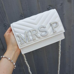 May include: White quilted clutch bag with a pearl embellished "MRS P" design. The bag has a pearl chain strap.