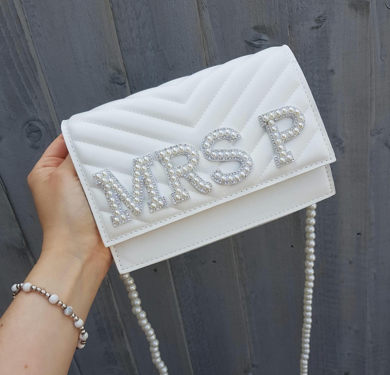 May include: White quilted clutch bag with a pearl embellished "MRS P" design. The bag has a pearl chain strap.