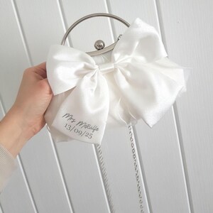 May include: A white satin clutch bag with a large white satin bow. The bag has a silver chain strap and a silver clasp. The bag is personalized with the text 'Mrs. Metcalfe 13/09/25'.