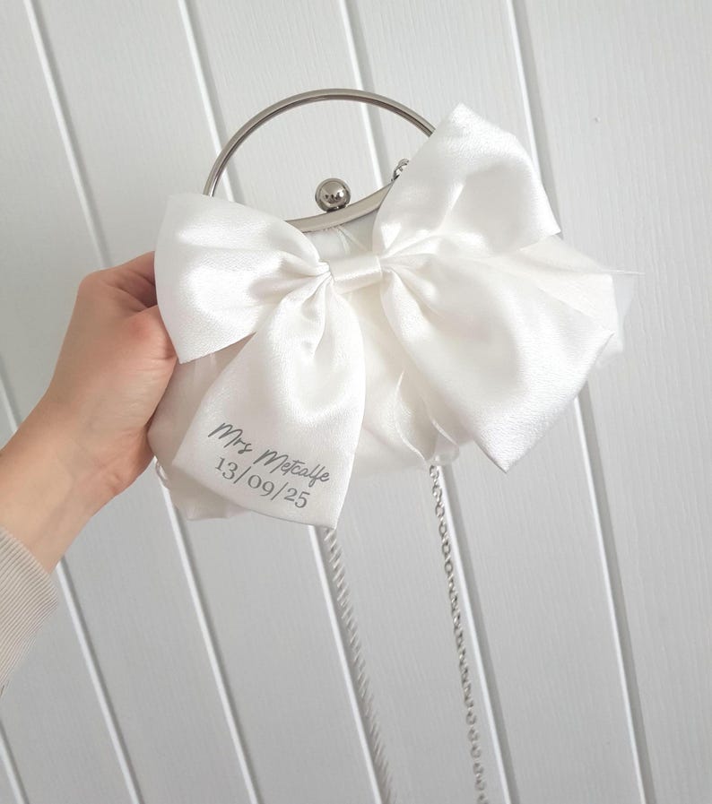 May include: A white satin clutch bag with a large white satin bow. The bag has a silver chain strap and a silver clasp. The bag is personalized with the text 'Mrs. Metcalfe 13/09/25'.