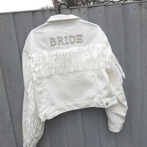 May include: White denim jacket with fringe detailing and the word "BRIDE" spelled out in pearl-like embellishments across the back. The jacket has a classic collar, button closures, and long sleeves. A stylish bridal jacket.