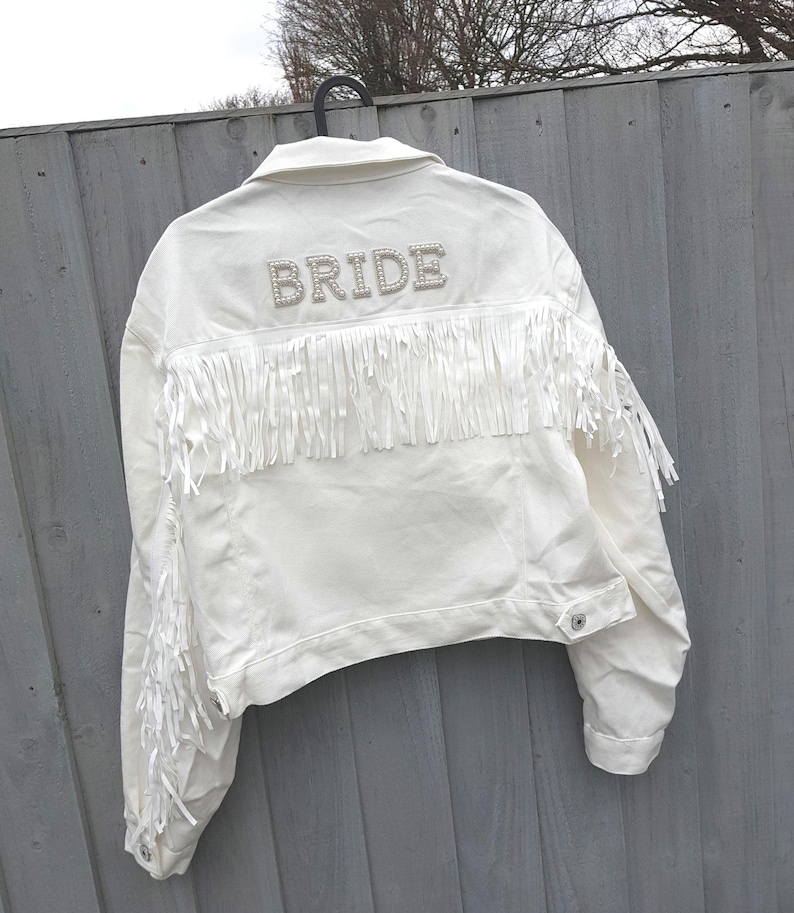 May include: White denim jacket with fringe detailing and the word "BRIDE" spelled out in pearl-like embellishments across the back. The jacket has a classic collar, button closures, and long sleeves. A stylish bridal jacket.