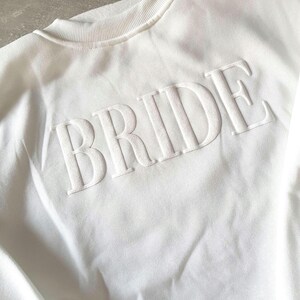 May include: A white crew neck sweatshirt with the word "BRIDE" in raised, textured lettering across the chest. The sweatshirt has a ribbed collar and a soft, casual appearance, perfect for bridal wear.