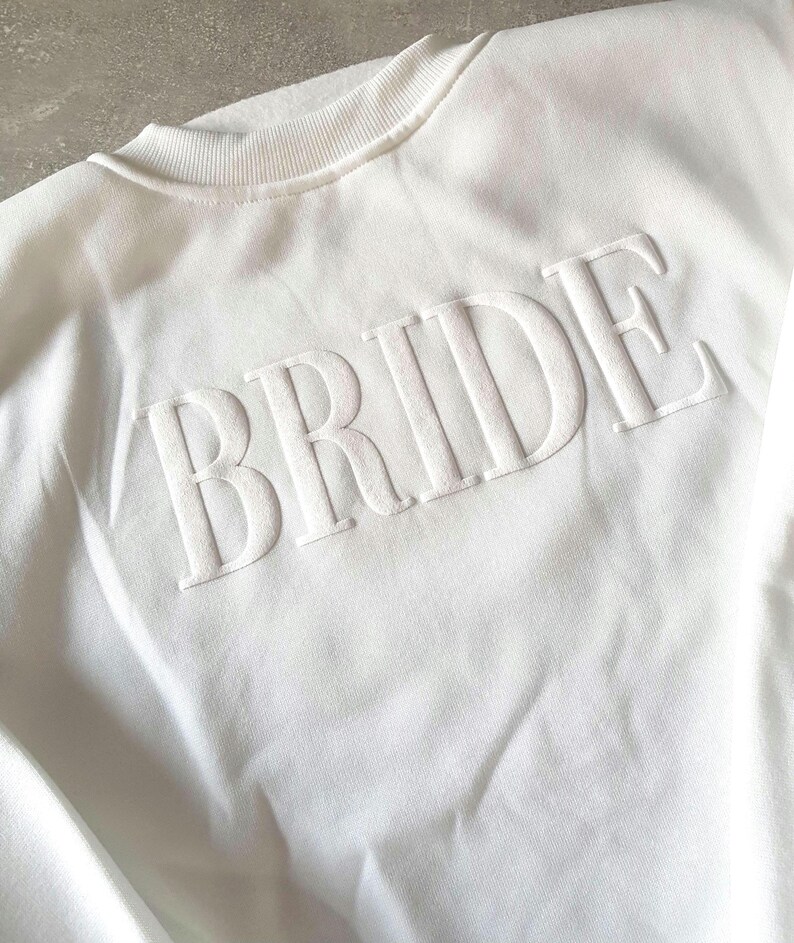 May include: A white crew neck sweatshirt with the word "BRIDE" in raised, textured lettering across the chest. The sweatshirt has a ribbed collar and a soft, casual appearance, perfect for bridal wear.