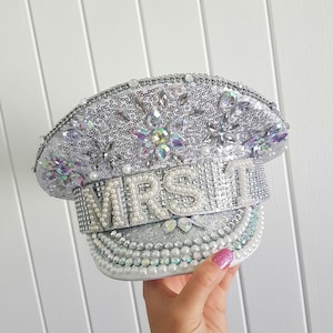 Op de afbeelding: Zilveren kapiteinshoed met pailletten met het woord "MRS" uitgespeld in grote parels. De hoed is versierd met iriserende strass-steentjes en parels. De rand is ook versierd met parels en strass-steentjes.