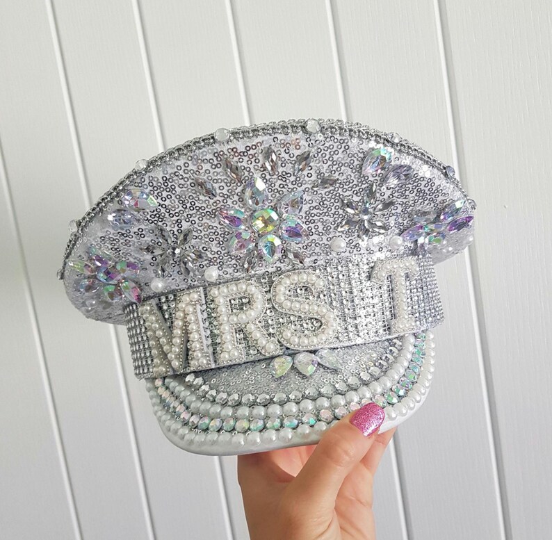 Op de afbeelding: Zilveren kapiteinshoed met pailletten met het woord "MRS" uitgespeld in grote parels. De hoed is versierd met iriserende strass-steentjes en parels. De rand is ook versierd met parels en strass-steentjes.