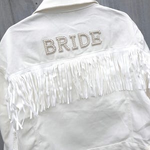 May include: White denim jacket with the word "BRIDE" spelled out in pearl-like beads across the back. The jacket features long white fringe across the upper back. A stylish bridal jacket.