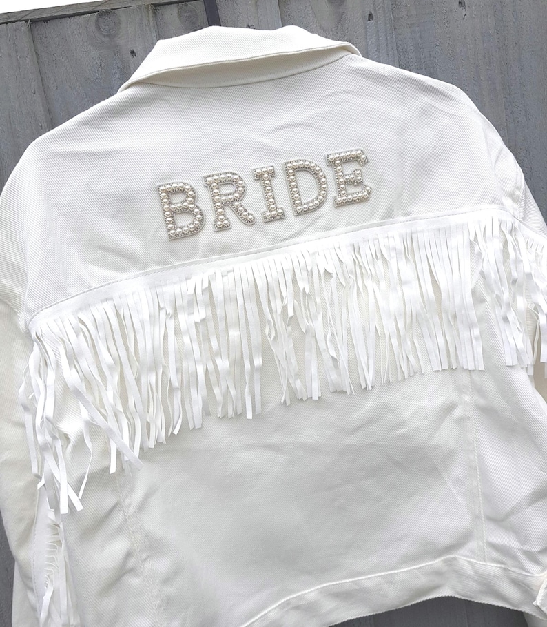 May include: White denim jacket with the word "BRIDE" spelled out in pearl-like beads across the back. The jacket features long white fringe across the upper back. A stylish bridal jacket.