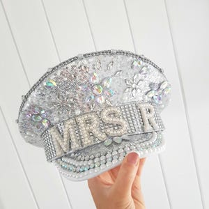 Op de afbeelding: Zilveren pailletten hoed met het woord "MRS" in parel- en strassletters. De hoed is versierd met iriserende edelstenen en parels. De hoed is een bruidsaccessoire.