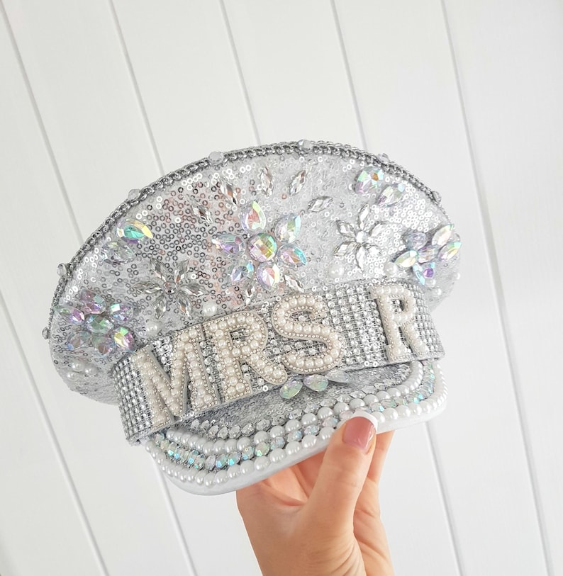 Op de afbeelding: Zilveren pailletten hoed met het woord "MRS" in parel- en strassletters. De hoed is versierd met iriserende edelstenen en parels. De hoed is een bruidsaccessoire.