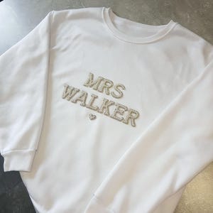 May include: White sweatshirt with the text "MRS WALKER" in silver rhinestones. A small heart is below the name. The sweatshirt has a crew neck and long sleeves with ribbed cuffs.