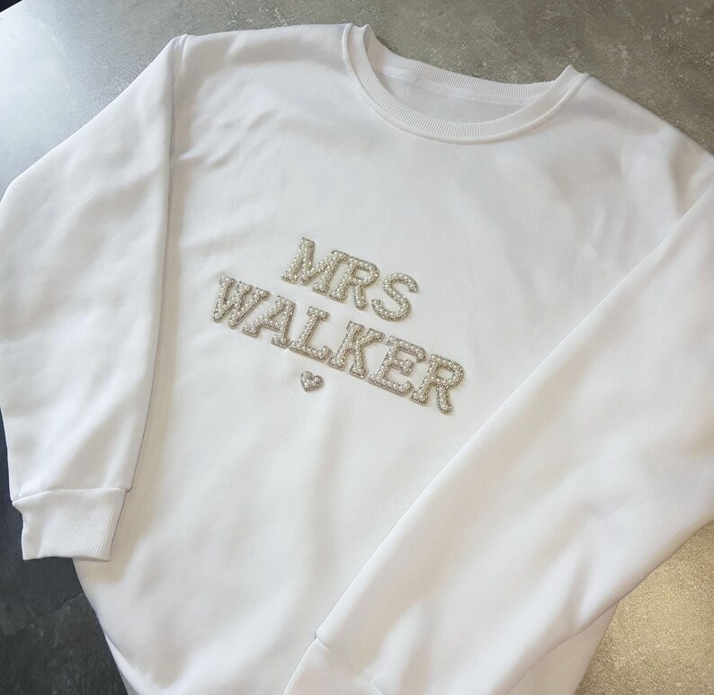 May include: White sweatshirt with the text "MRS WALKER" in silver rhinestones. A small heart is below the name. The sweatshirt has a crew neck and long sleeves with ribbed cuffs.