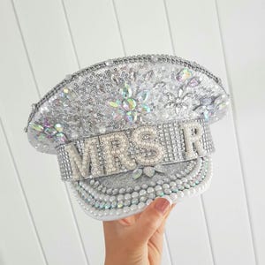 Op de afbeelding: Een zilveren pailletten hoed met het woord "MRS R" in parelachtige letters. De hoed is versierd met iriserende edelstenen en witte parels. De hoed is een bruidsaccessoire.
