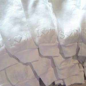 May include: Close-up of several white sweatshirts with embroidered initials and heart symbols on the sleeves. The initials include "L J", "L", "S D", and "K & S". The sweatshirts are folded and stacked.