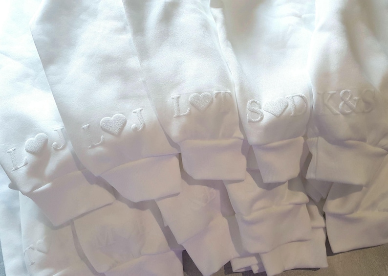 May include: Close-up of several white sweatshirts with embroidered initials and heart symbols on the sleeves. The initials include "L J", "L", "S D", and "K & S". The sweatshirts are folded and stacked.