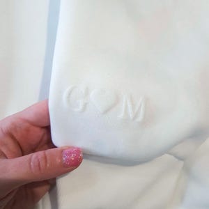 May include: Close-up of a white sweatshirt sleeve. The sleeve features embossed initials "G" and "M" separated by a heart symbol. The fabric appears soft and the design is subtle. The overall aesthetic is minimalist and personalized.