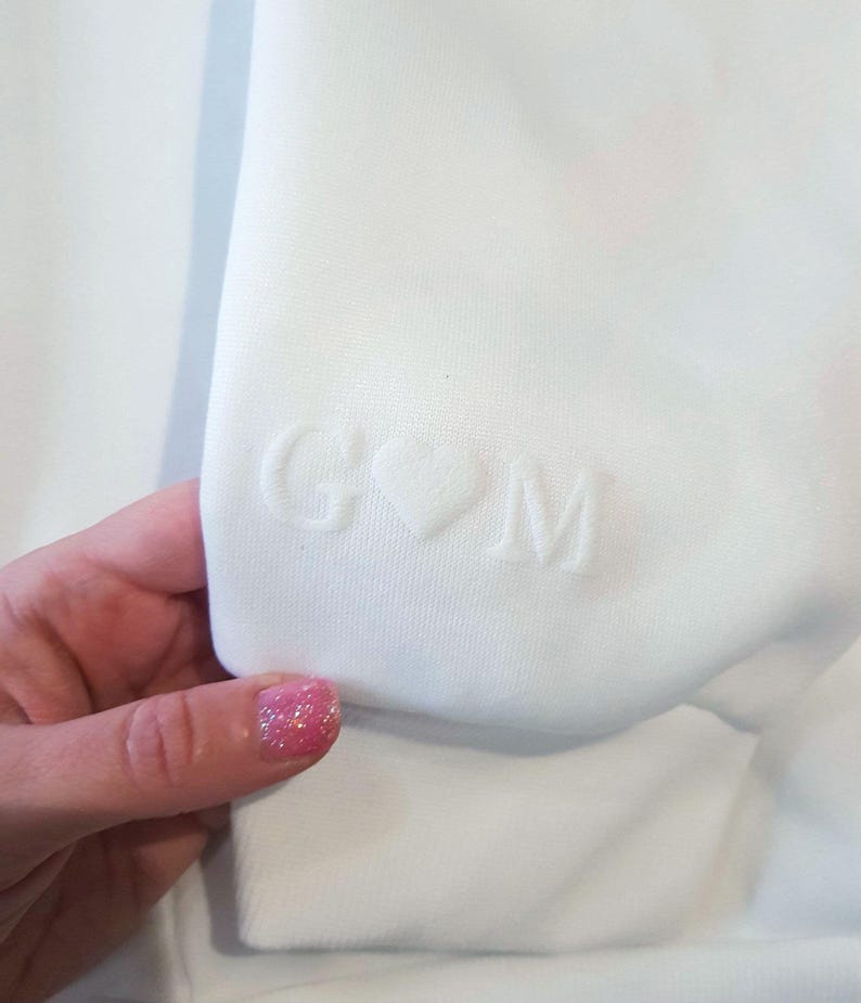 May include: Close-up of a white sweatshirt sleeve. The sleeve features embossed initials "G" and "M" separated by a heart symbol. The fabric appears soft and the design is subtle. The overall aesthetic is minimalist and personalized.