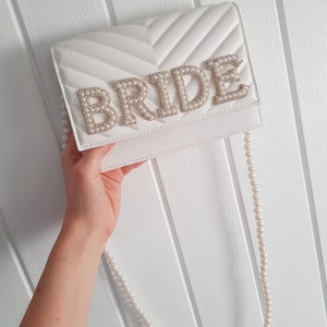 May include: White quilted clutch bag with a pearl embellished "BRIDE" detail and a matching pearl chain strap.
