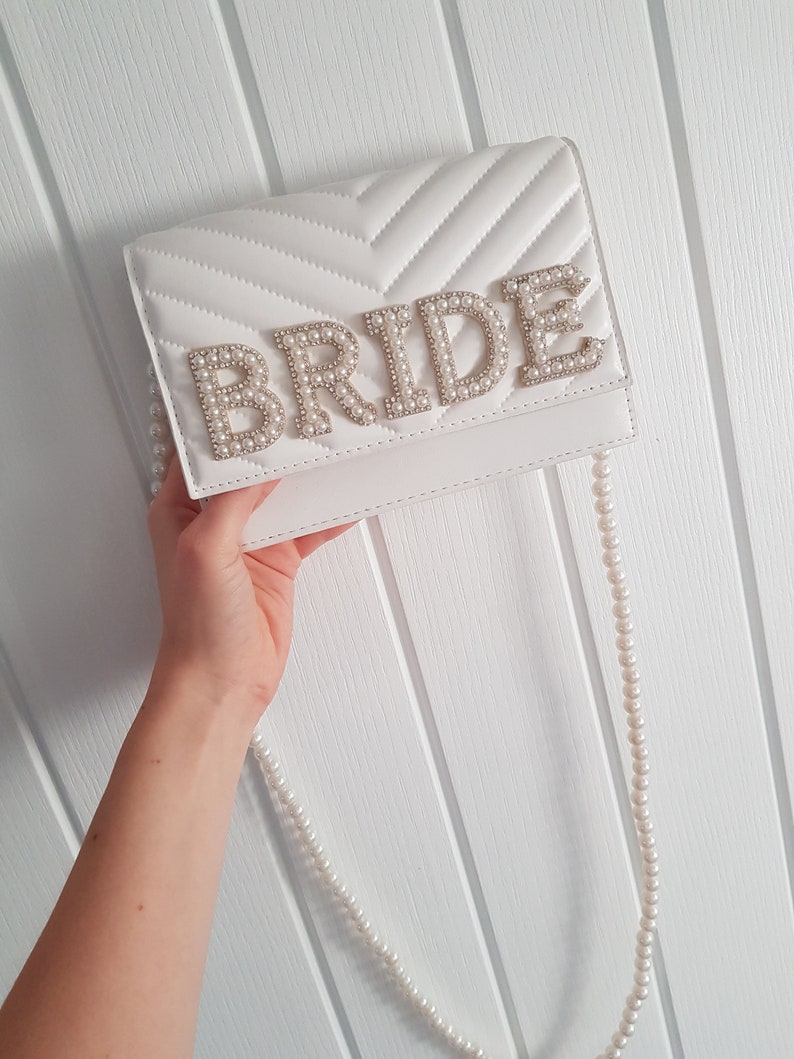 May include: White quilted clutch bag with a pearl embellished "BRIDE" detail and a matching pearl chain strap.