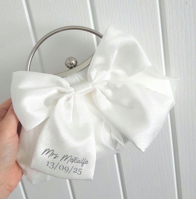 May include: White satin clutch bag with a large bow and a silver handle. The bag has the text "Mrs. Metcalfe 13/09/25" printed on it.
