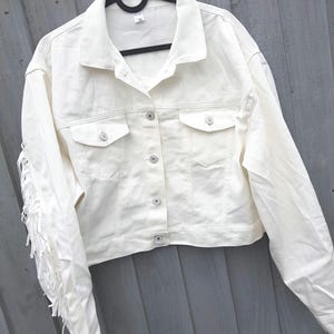 May include: Off-white denim jacket with fringe detail on the sleeves. The jacket features a classic collar, button-down closure, and two chest pockets. The jacket is hanging on a black hanger.
