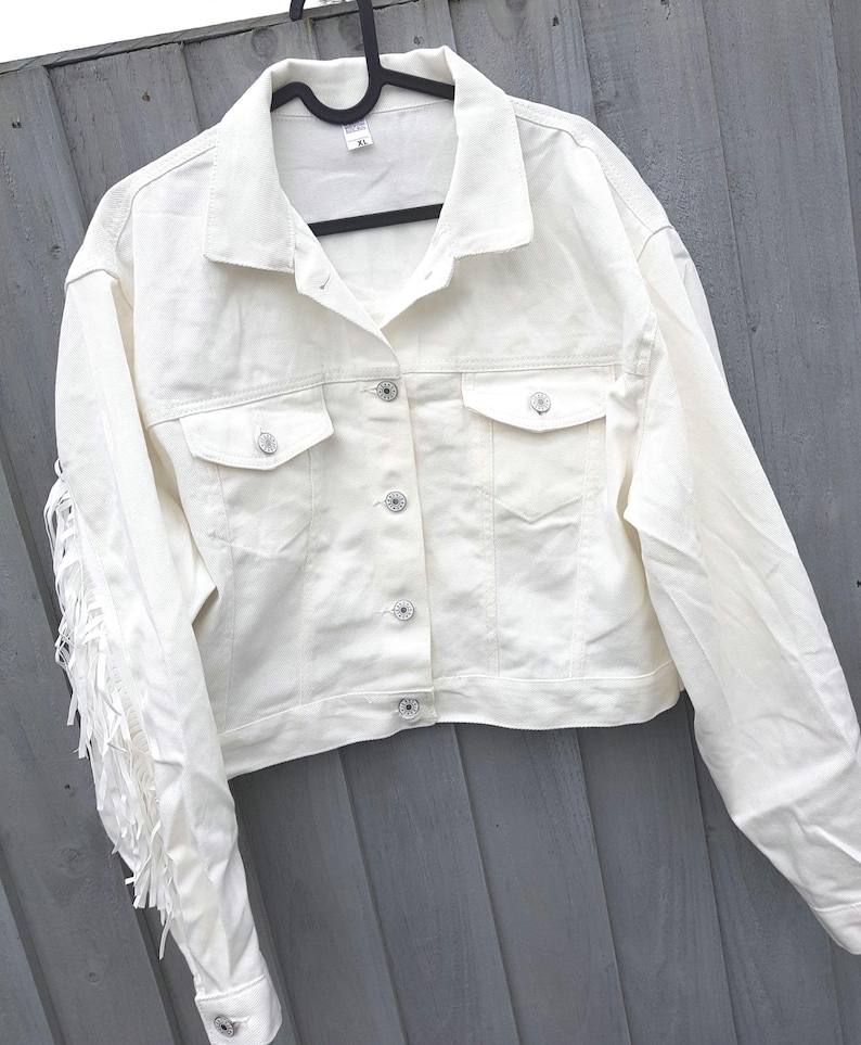 May include: Off-white denim jacket with fringe detail on the sleeves. The jacket features a classic collar, button-down closure, and two chest pockets. The jacket is hanging on a black hanger.