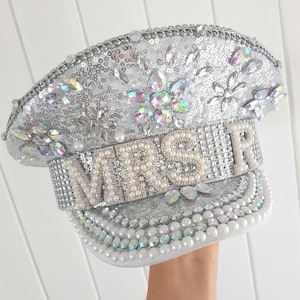 Op de afbeelding: Zilveren pailletten hoed met het woord "MRS" in parelachtige kralen. De hoed is versierd met iriserende strass en parels. De rand en de kroon zijn rijkelijk versierd met pailletten en edelstenen, wat een glamoureus accessoire cre&euml;ert.