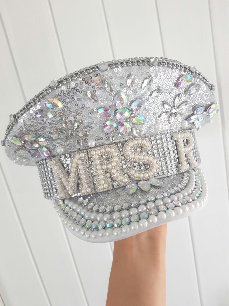 Op de afbeelding: Zilveren pailletten hoed met het woord "MRS" in parelachtige kralen. De hoed is versierd met iriserende strass en parels. De rand en de kroon zijn rijkelijk versierd met pailletten en edelstenen, wat een glamoureus accessoire cre&euml;ert.