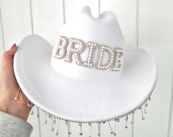 May include: White cowboy hat with the word "BRIDE" spelled out in pearl-like letters. The hat has a silver beaded trim and dangling silver accents around the brim. A festive accessory for a bachelorette party or wedding.