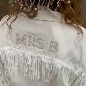 May include: White jacket with fringe detail and the text "MRS B" in sparkling embellishments. The jacket has a decorative headpiece with dangling rhinestones. A bridal or special occasion garment.