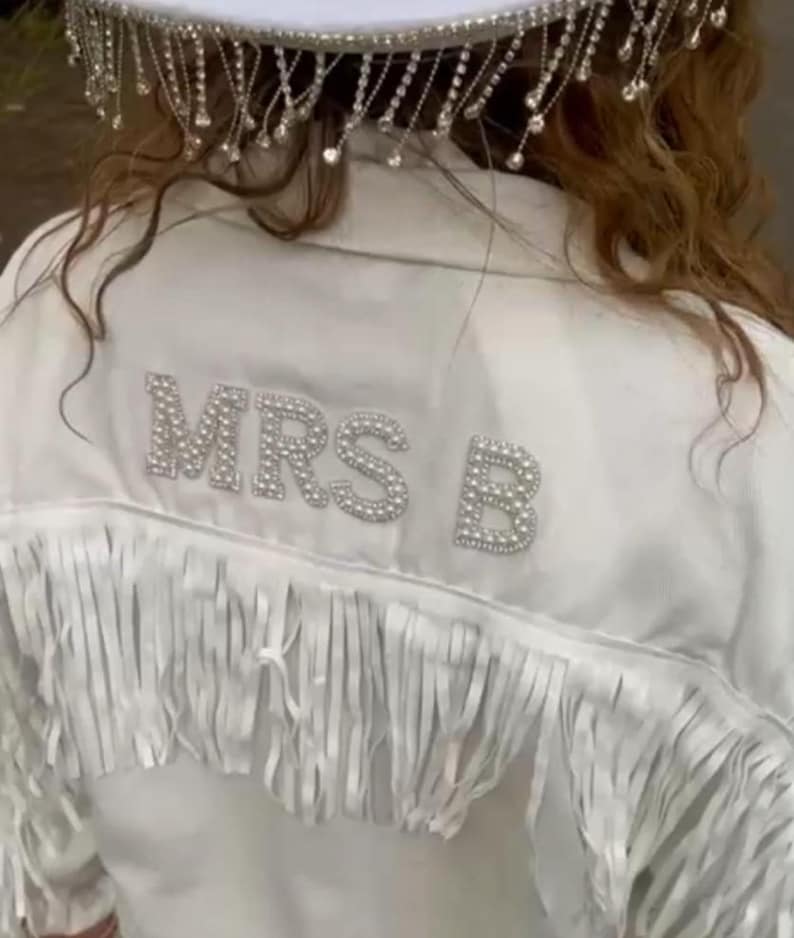 May include: White jacket with fringe detail and the text "MRS B" in sparkling embellishments. The jacket has a decorative headpiece with dangling rhinestones. A bridal or special occasion garment.