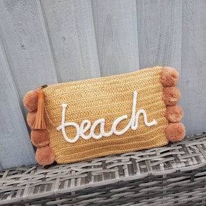 May include: A straw beach bag with a white "beach" embroidered on the front. The bag has brown pom-poms around the edges.