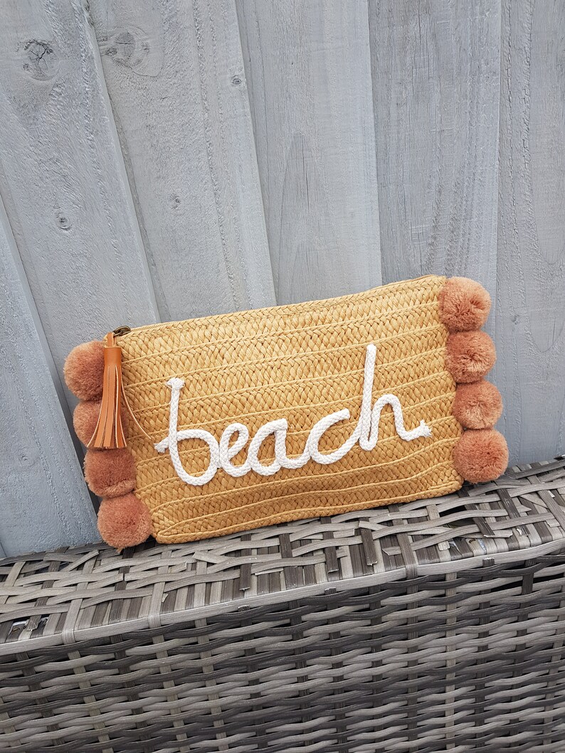 May include: A straw beach bag with a white "beach" embroidered on the front. The bag has brown pom-poms around the edges.