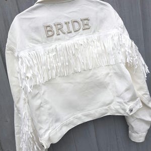 May include: White denim jacket with fringe detailing and the word "BRIDE" spelled out in pearl-like beads across the back. The jacket has a classic collar and button closures. The fringe is along the back and sleeves.