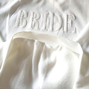 May include: White crew neck sweatshirt with the word "BRIDE" in raised, textured lettering across the chest. The sweatshirt has a soft, fleecy interior and a ribbed neckline and cuffs. The overall style is casual and comfortable.