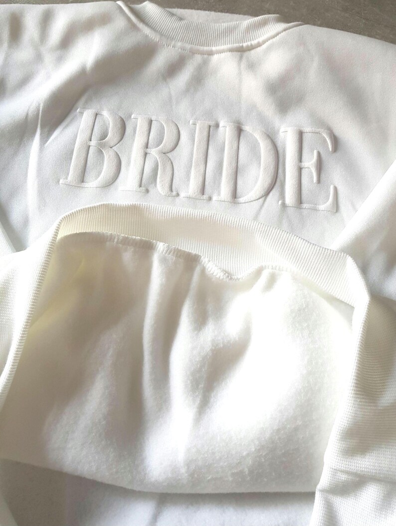 May include: White crew neck sweatshirt with the word "BRIDE" in raised, textured lettering across the chest. The sweatshirt has a soft, fleecy interior and a ribbed neckline and cuffs. The overall style is casual and comfortable.