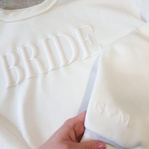 May include: White sweatshirt with the word "BRIDE" embossed across the chest. The sleeve features the initials "G♡M" embossed. The sweatshirt is a crew neck style and appears to be made of a soft material.