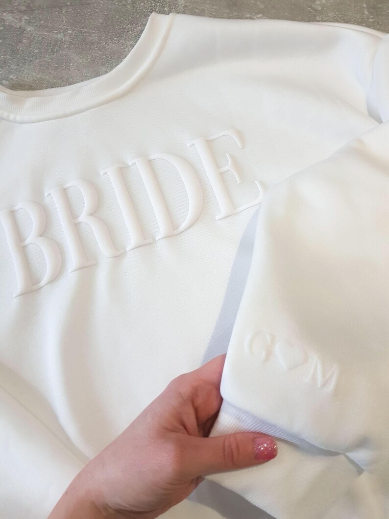 May include: White sweatshirt with the word "BRIDE" embossed across the chest. The sleeve features the initials "G♡M" embossed. The sweatshirt is a crew neck style and appears to be made of a soft material.