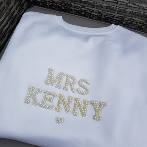 May include: White crew neck sweatshirt with the text "MRS KENNY" in pearl-like embellishments. A small heart-shaped embellishment is below the name. The sweatshirt is made of a soft material.