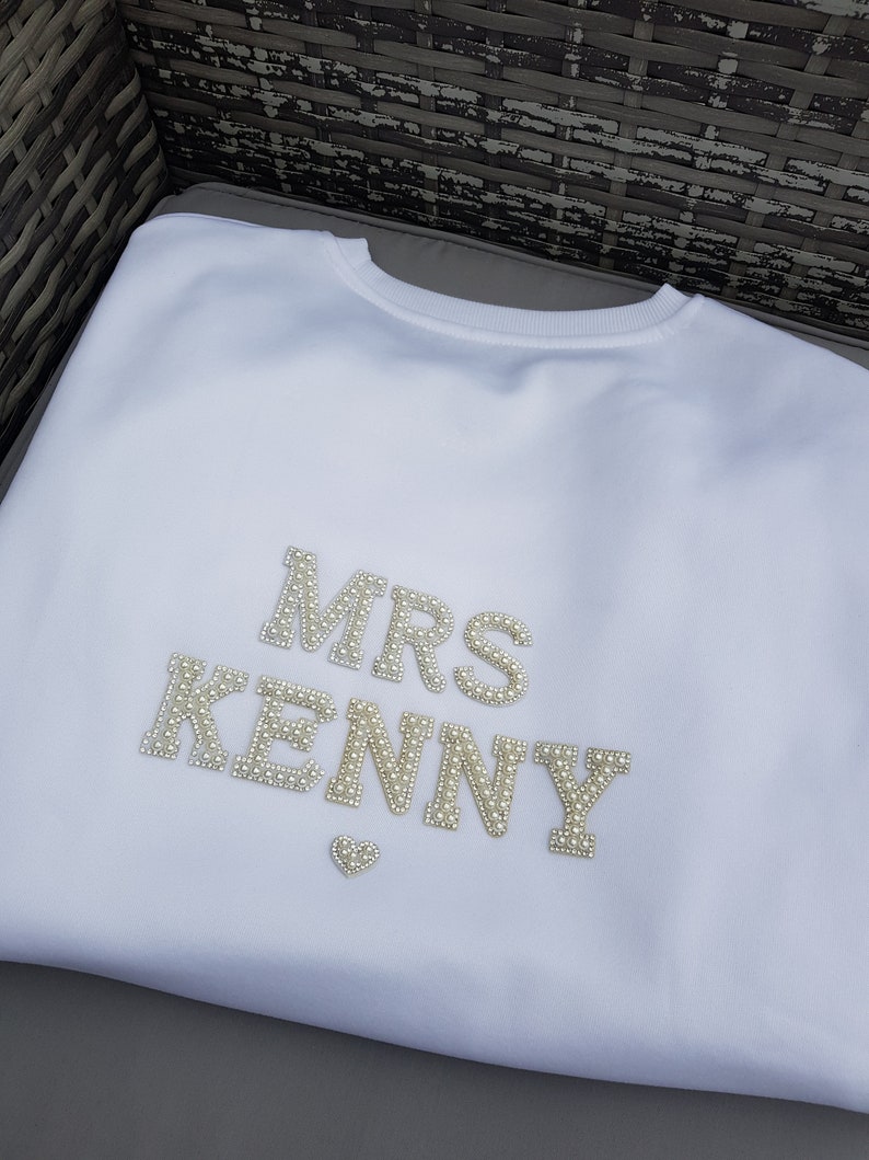 May include: White crew neck sweatshirt with the text "MRS KENNY" in pearl-like embellishments. A small heart-shaped embellishment is below the name. The sweatshirt is made of a soft material.