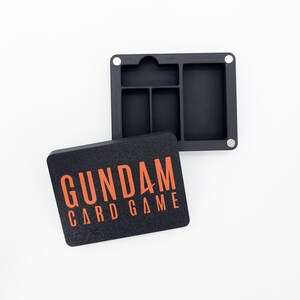 Puede incluir: Caja negra para juego de cartas con las palabras "GUNDAM CARD GAME" en naranja. La caja está abierta, revelando compartimentos para organizar los componentes del juego. La caja es cuadrada con esquinas redondeadas.