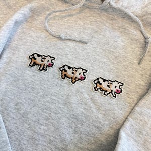 May include: A light gray heathered hoodie featuring three embroidered pixel art cows arranged in a horizontal row. Each cow is rendered in black, white, and tan thread with a small pink snout, showcasing a distinct blocky, retro-inspired design.