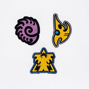 Op de afbeelding: Drie geborduurde patches met unieke ontwerpen. Eén is een paarse spiraal op een zwarte achtergrond, een andere is een gele vlamvorm met een blauwe stip, en de derde is een gele stervorm met een blauwe rand.