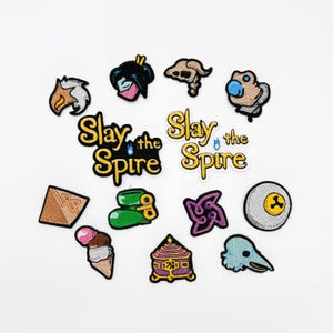 May include: A selection of embroidered patches with diverse designs, such as an ice cream cone, a skull, and a stylized bird. The text "Slay the Spire" is displayed on two patches. The patches are in various colours, including black, gold, green, and purple.