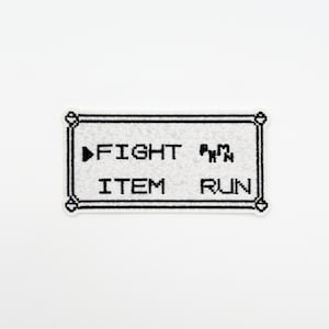 May include: A rectangular, off-white patch with black embroidered text and border. The text reads "FIGHT", "ITEM", and "RUN", with a black arrow pointing to "FIGHT". The patch has a pixelated aesthetic, reminiscent of a classic video game.